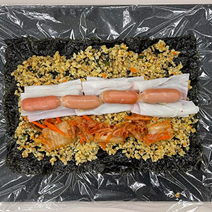 海苔底部鋪上保鮮膜，接續在海苔上放上蕎麥花椰菜飯、德式起司腸、泡菜、蟳味棒。